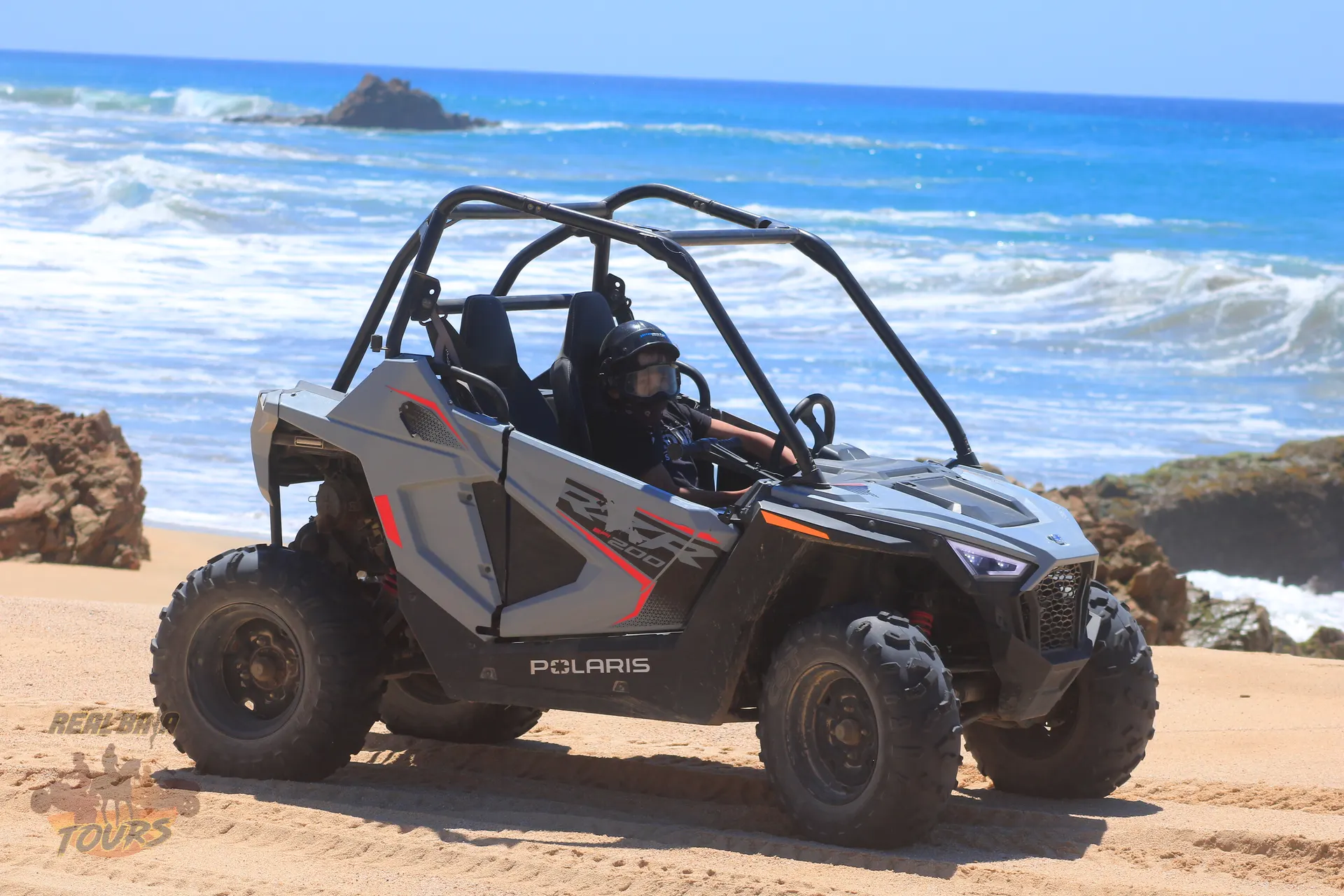 Polaris RZR side-by-side vehicle parked on sandy beach with turquoise ocean waves and rocky coastline in Baja California Sur