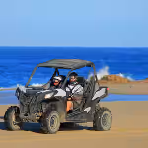 Two people riding a side-by-side UTV on a sandy beach with ocean waves and rocky outcrops in Baja California Sur