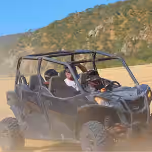 3 tourist riding in a black open-air UTV across dusty desert terrain with rocky hills in background near Los Cabos, Baja Cali